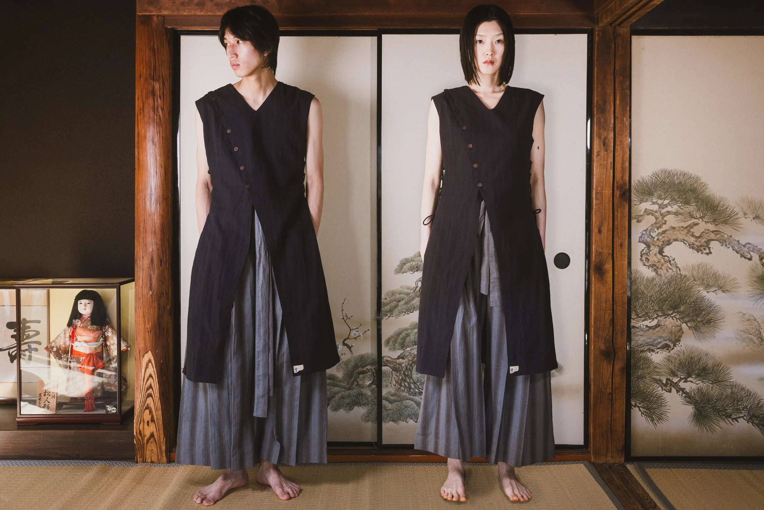 Person wearing a black sleeveless top and gray skirt standing in front of a wooden screen with traditional Japanese artwork.