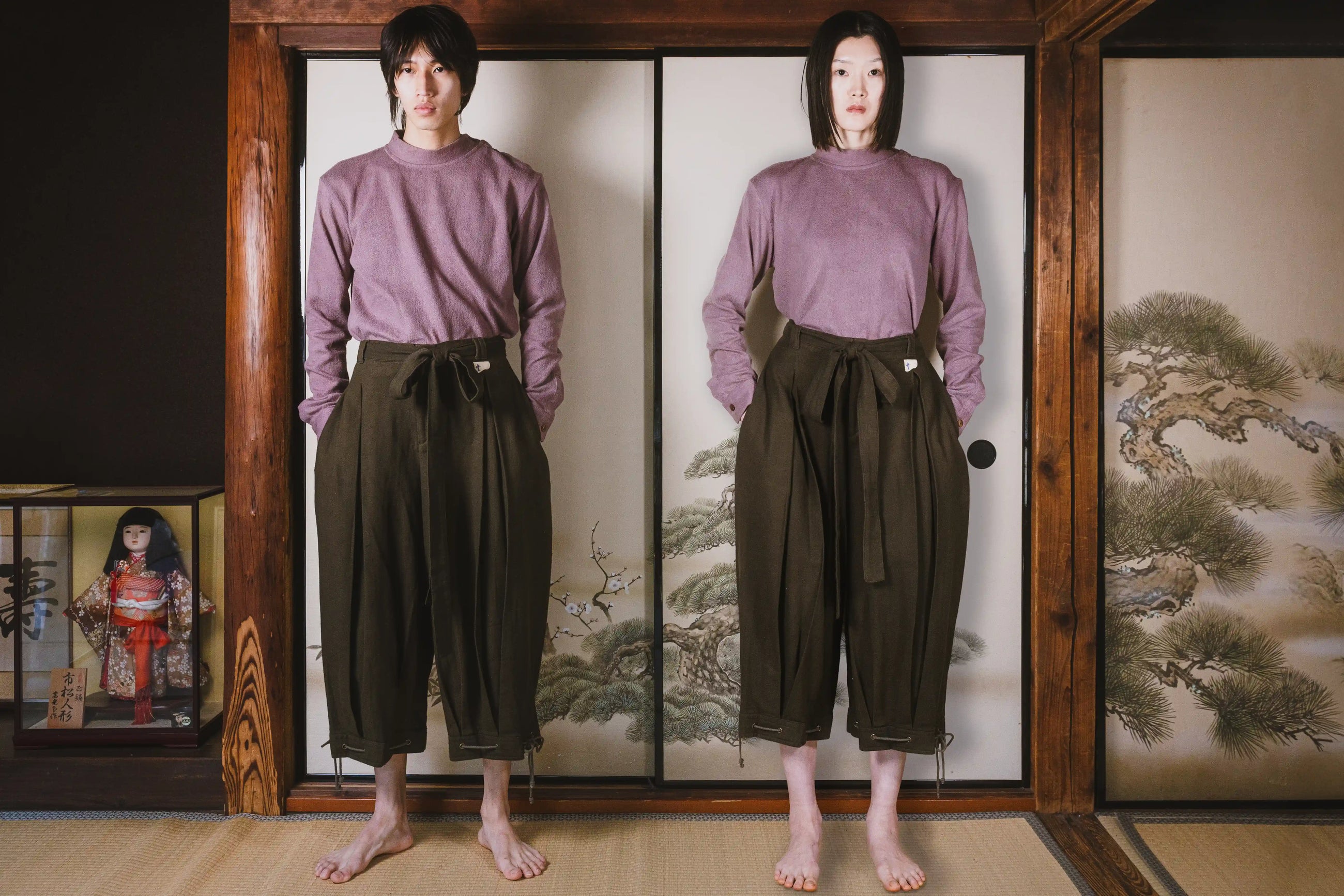 Person wearing a purple top and brown pants standing in front of a wooden screen with a painting.