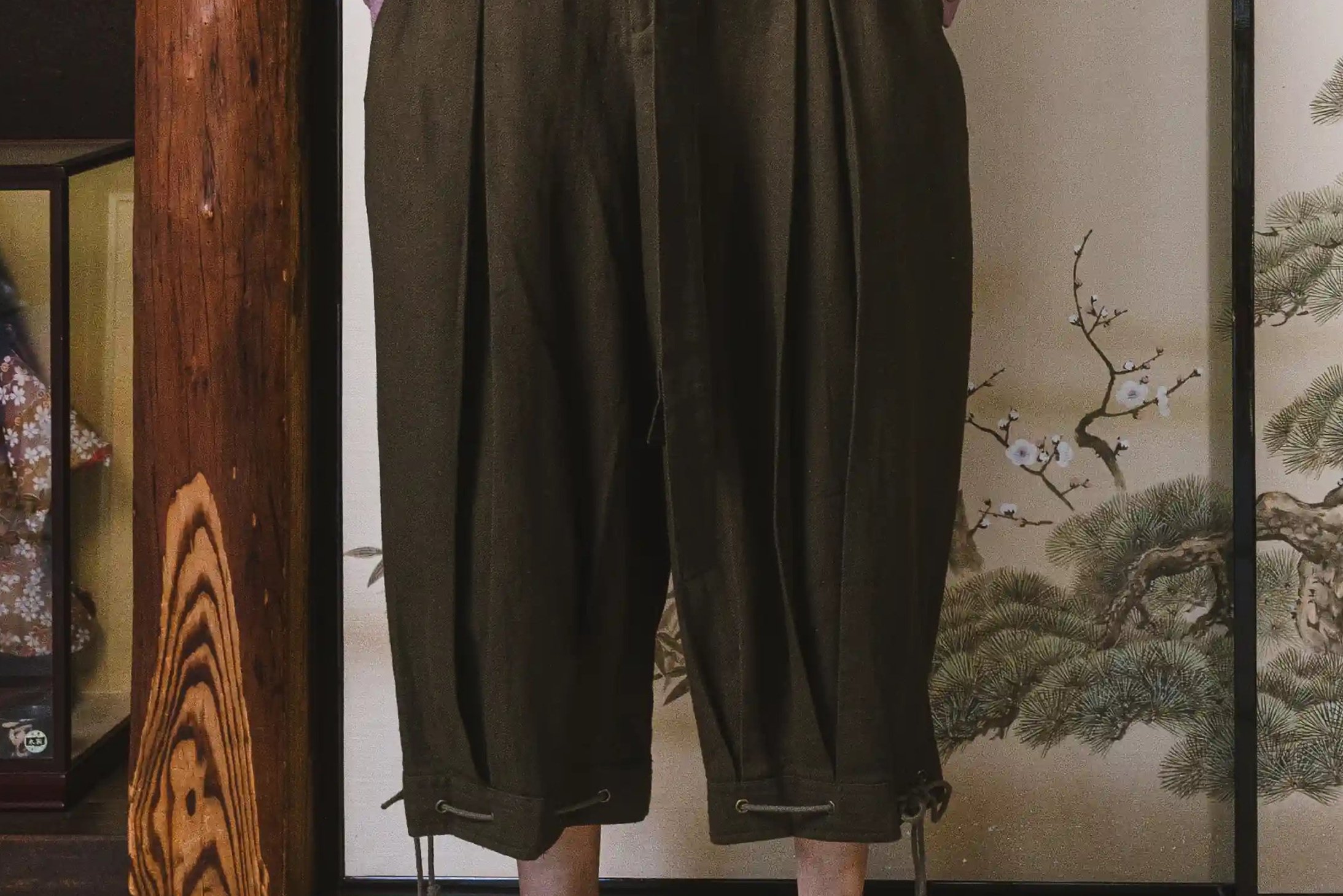 Person wearing dark green wide-leg pants standing in front of a decorative screen with floral patterns.