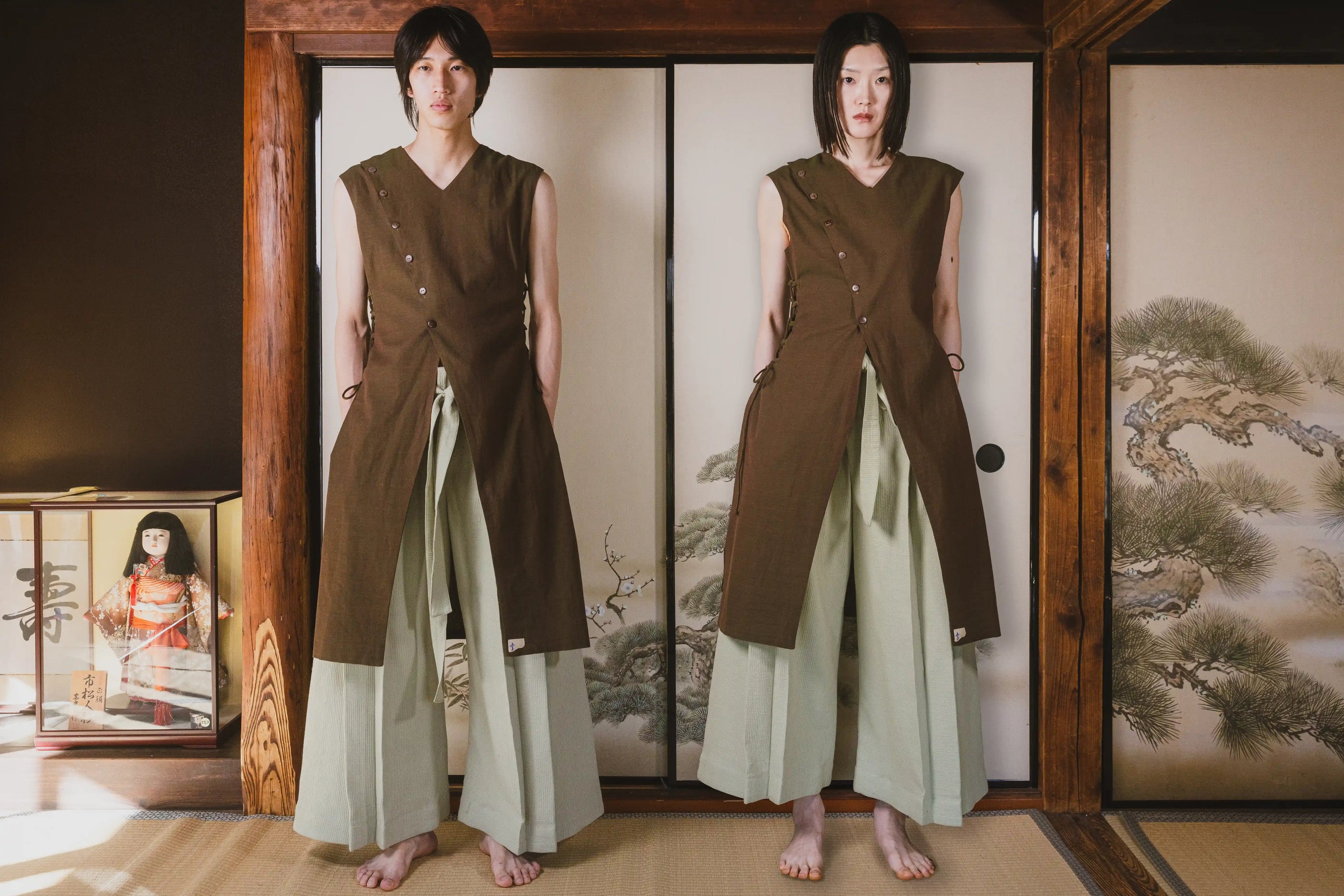 Person wearing a brown sleeveless top and light green pants standing in front of a wooden screen with traditional Japanese artwork.