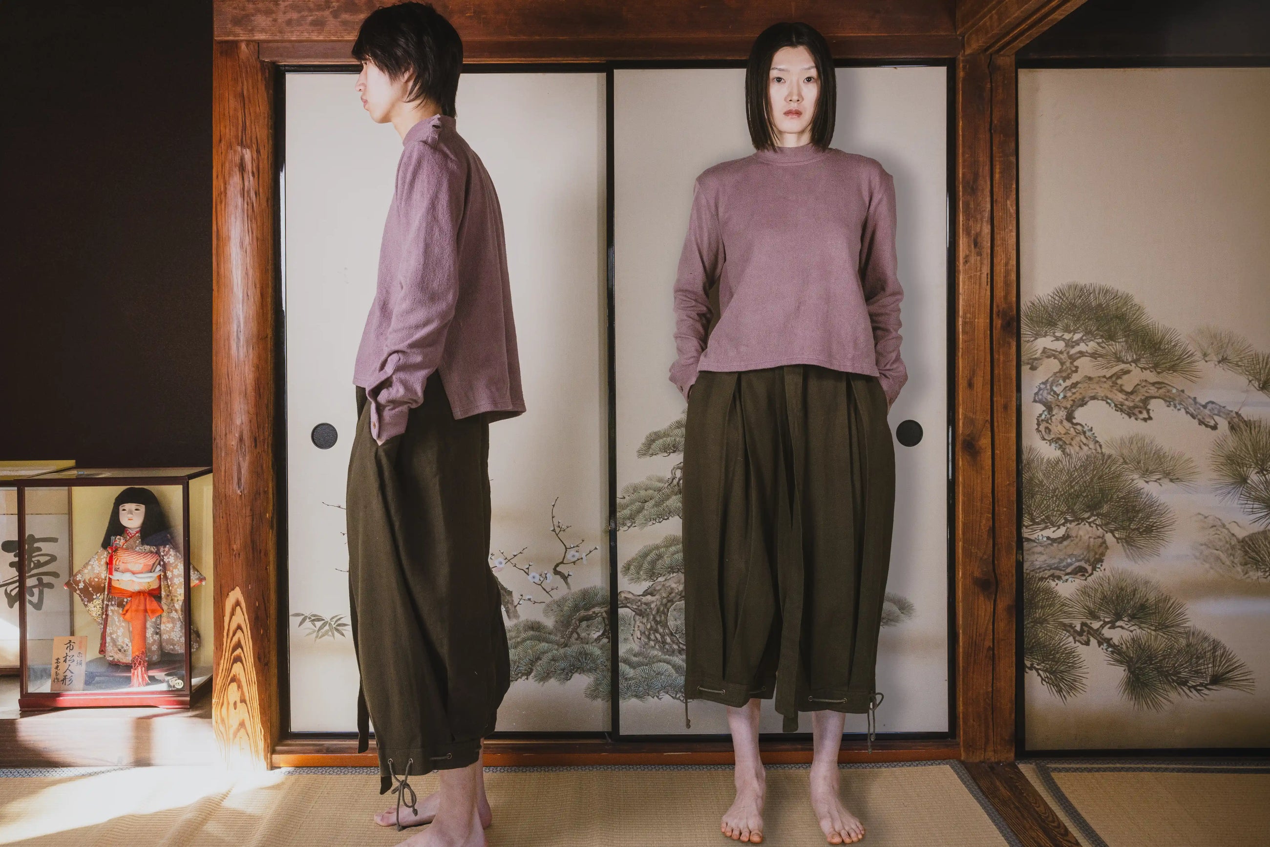 Person wearing a purple shirt and green pants standing in front of a traditional Japanese screen with a doll display.