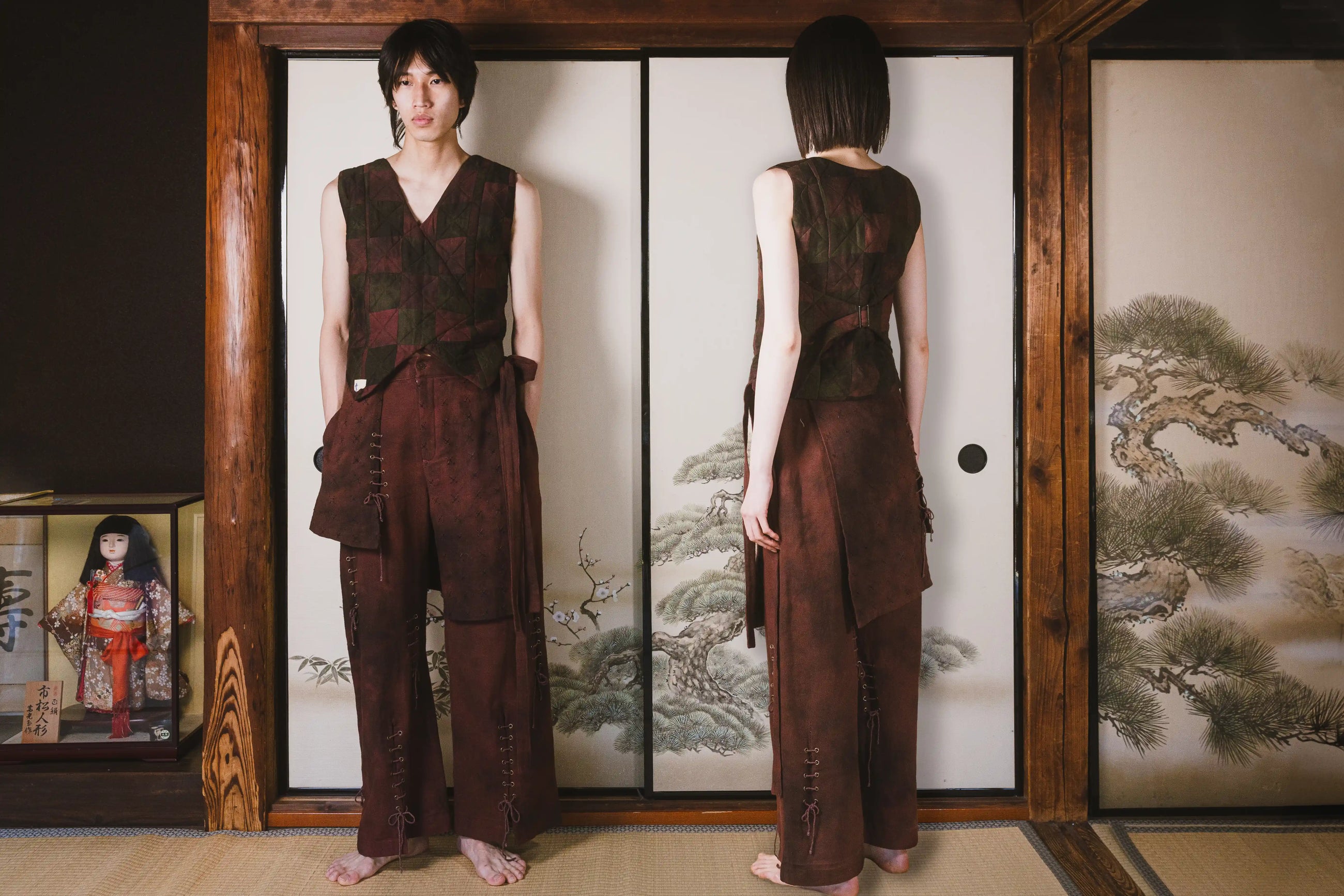 Person wearing a brown outfit standing in front of a traditional Japanese screen with a doll and artwork on the side.