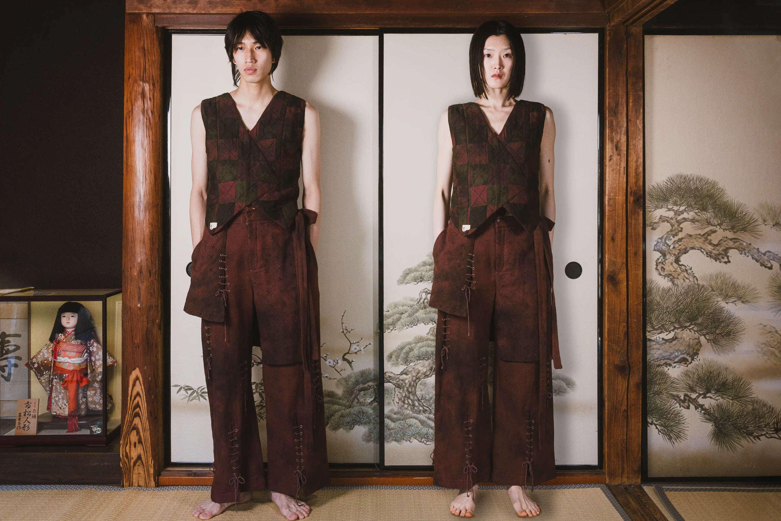 Two people wearing matching brown outfits standing in front of a traditional Japanese screen with nature scenes.