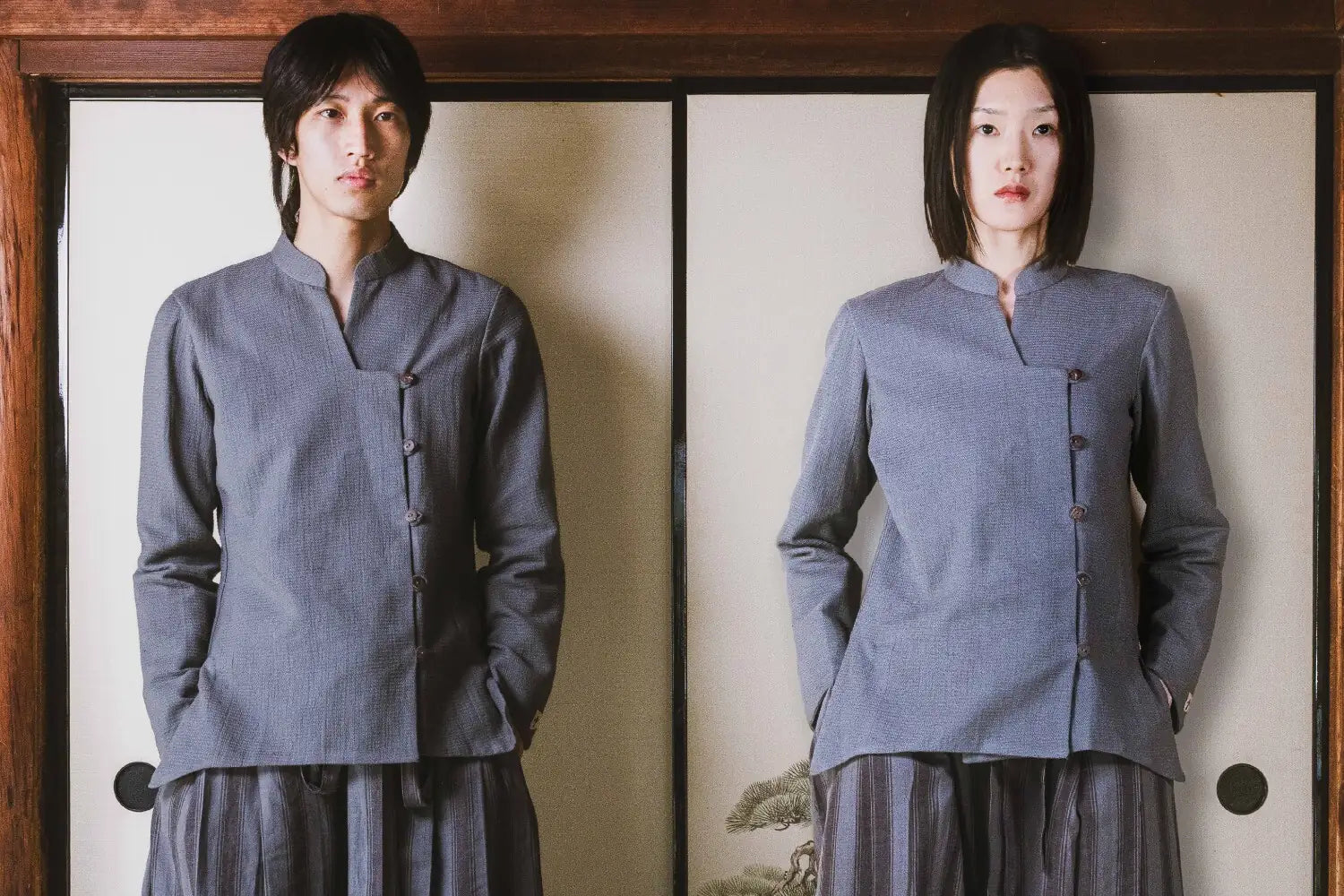 Two people wearing matching gray traditional outfits standing in front of a wooden frame.