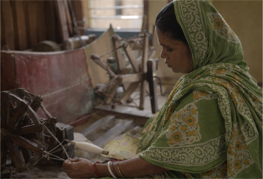 Woman spinning yarn using a traditional spinning wheel in a dimly lit room.