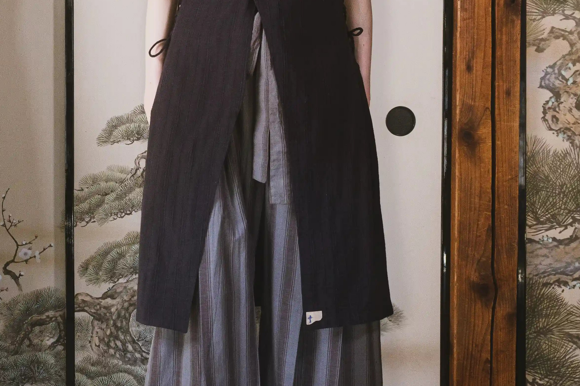 Person wearing a black vest over a light-colored dress in front of a traditional Japanese screen with nature designs.