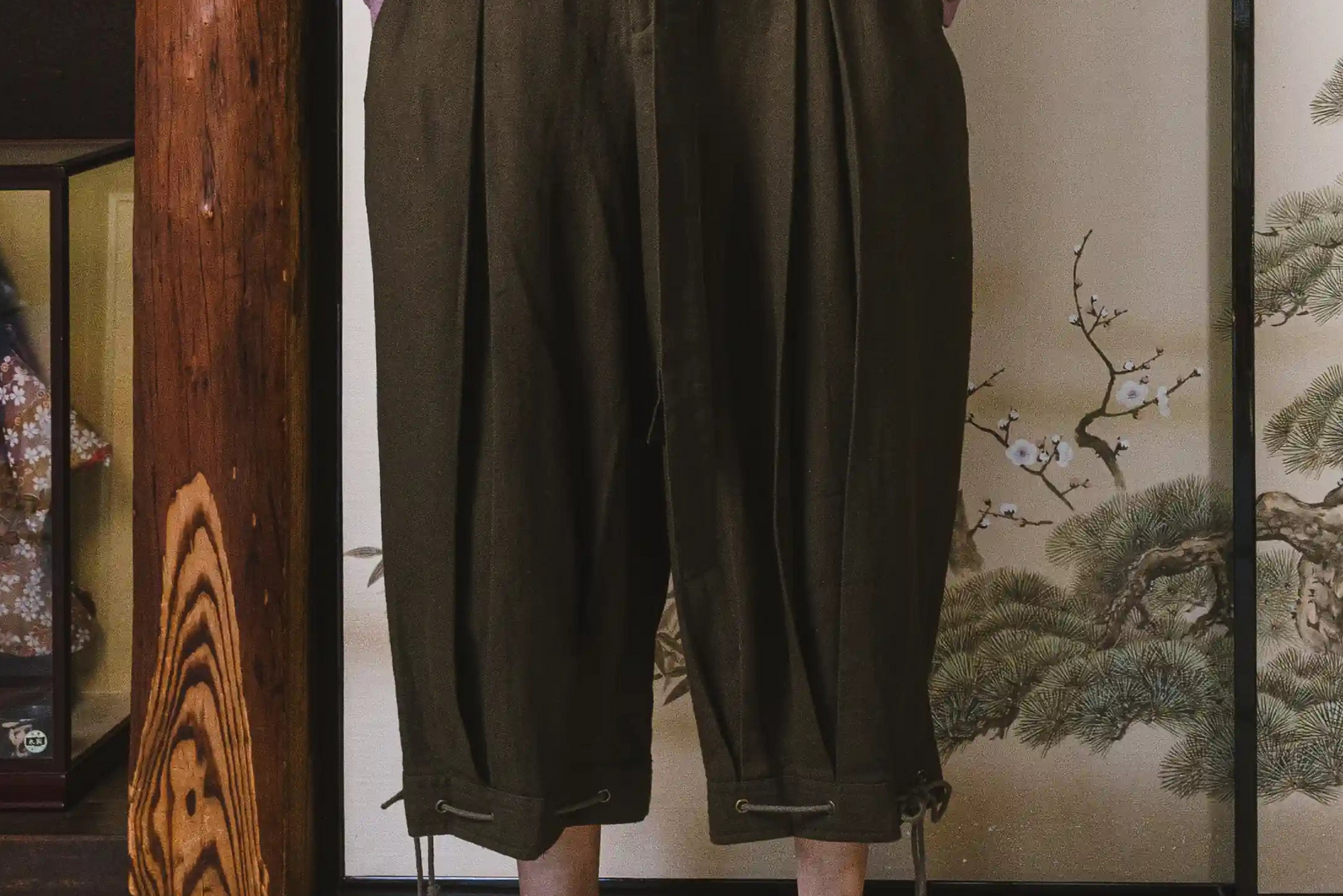 Person wearing dark green wide-leg pants standing in front of a decorative screen with floral patterns.