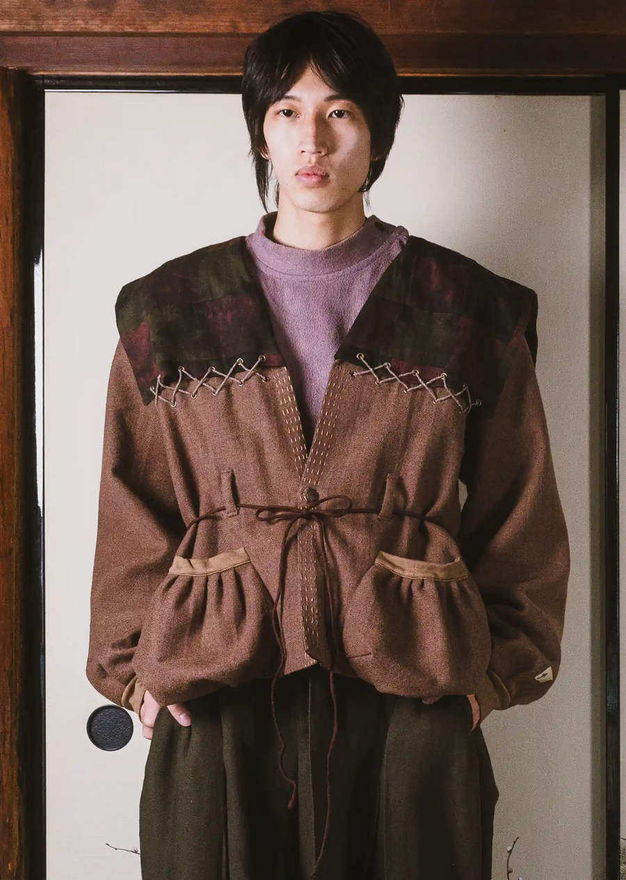 Person wearing a brown jacket with decorative stitching in front of a white wall.