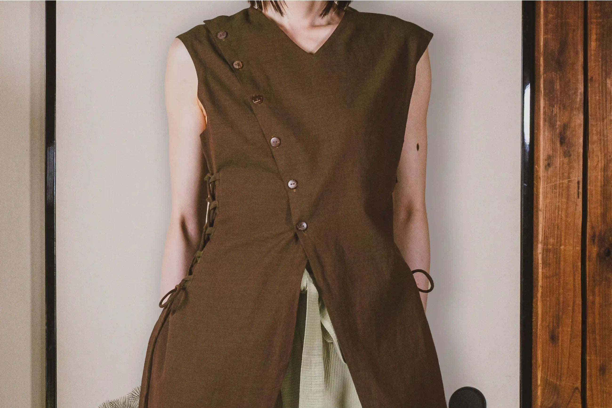Person wearing a brown sleeveless outfit with buttons against a neutral background