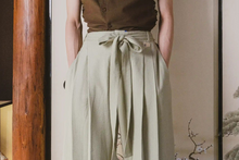 Person wearing a brown top and light green pleated pants against a neutral background