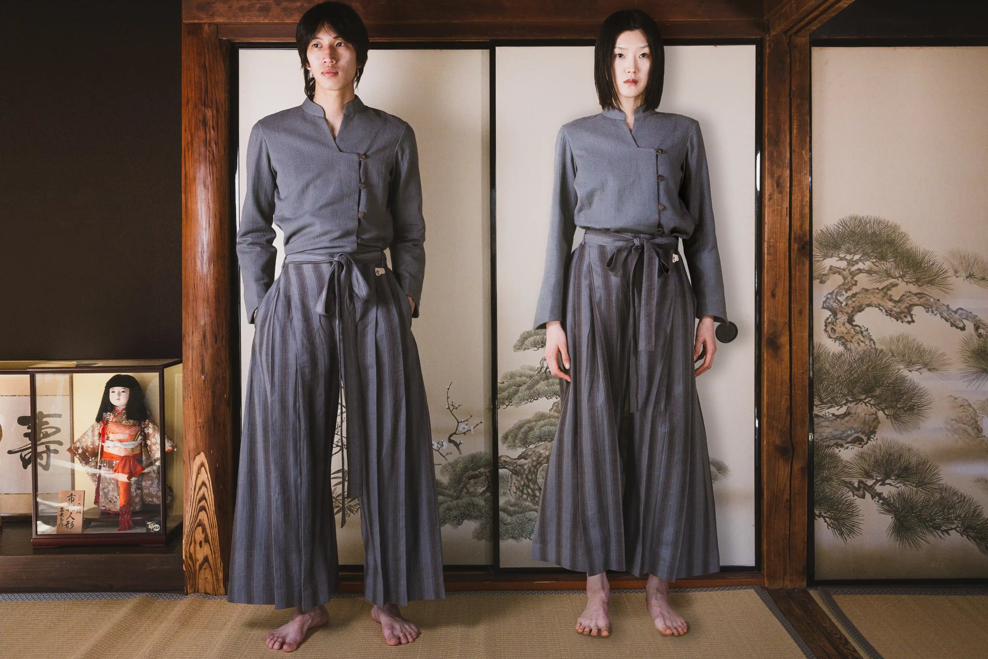 Person wearing a traditional gray outfit standing in front of a wooden screen with a landscape design.
