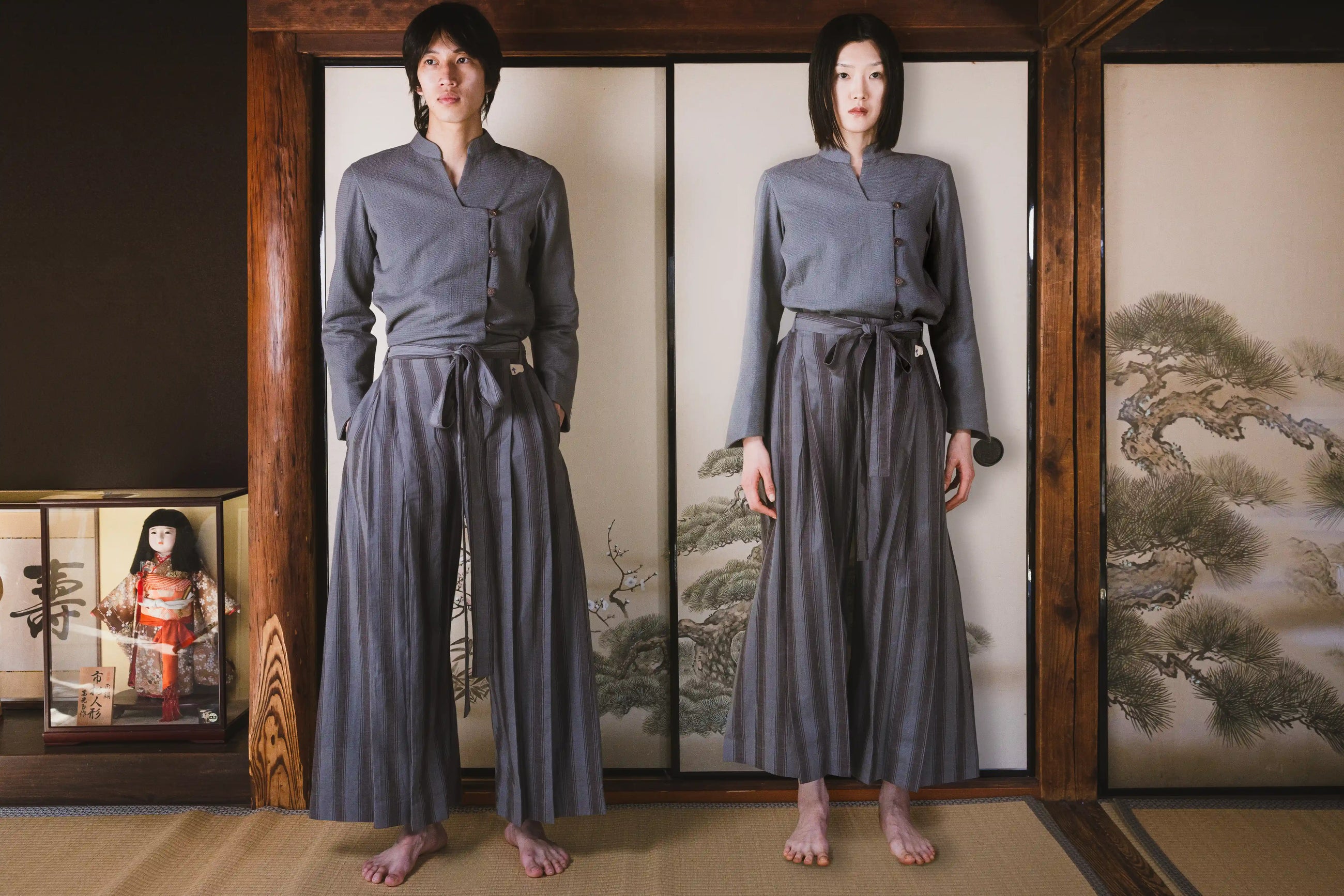 Person wearing a traditional gray outfit standing in front of a wooden screen with a landscape design.