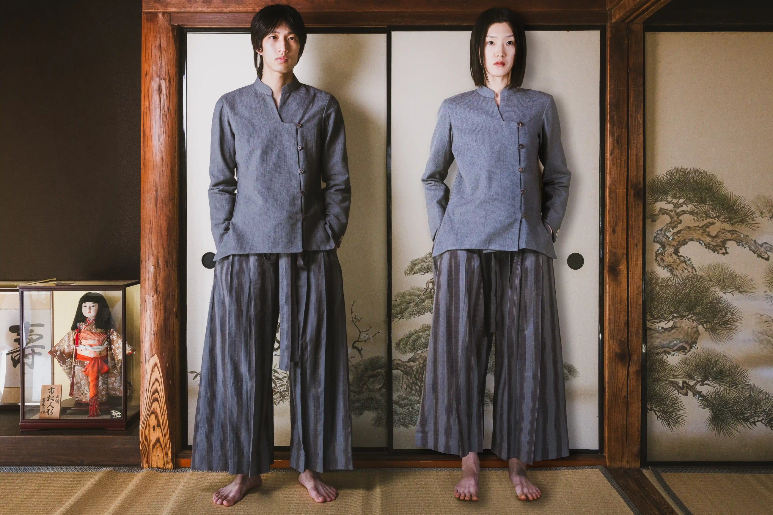 Two people wearing matching gray outfits standing in front of a wooden screen with traditional Japanese design.