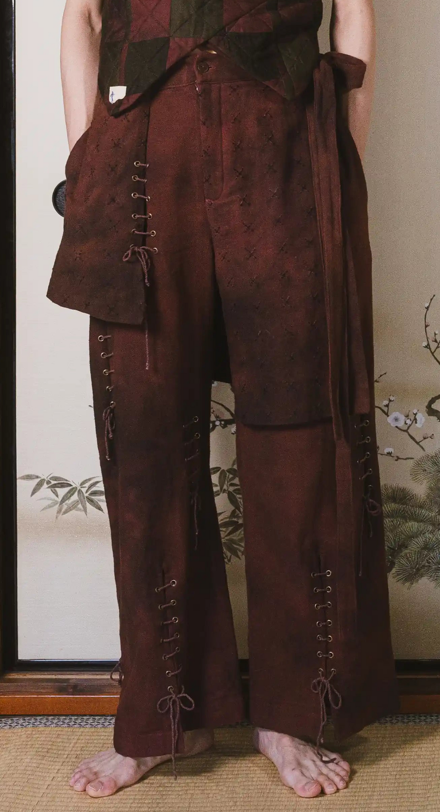 Person wearing a brown outfit with wide-leg pants and a jacket in front of a decorative wall.
