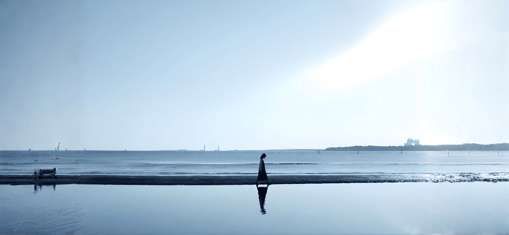 Person walking along a beach with a bright light source in the sky